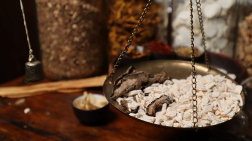 Close-up of traditional herbal medicine being weighed on a balance scale among various herbs.